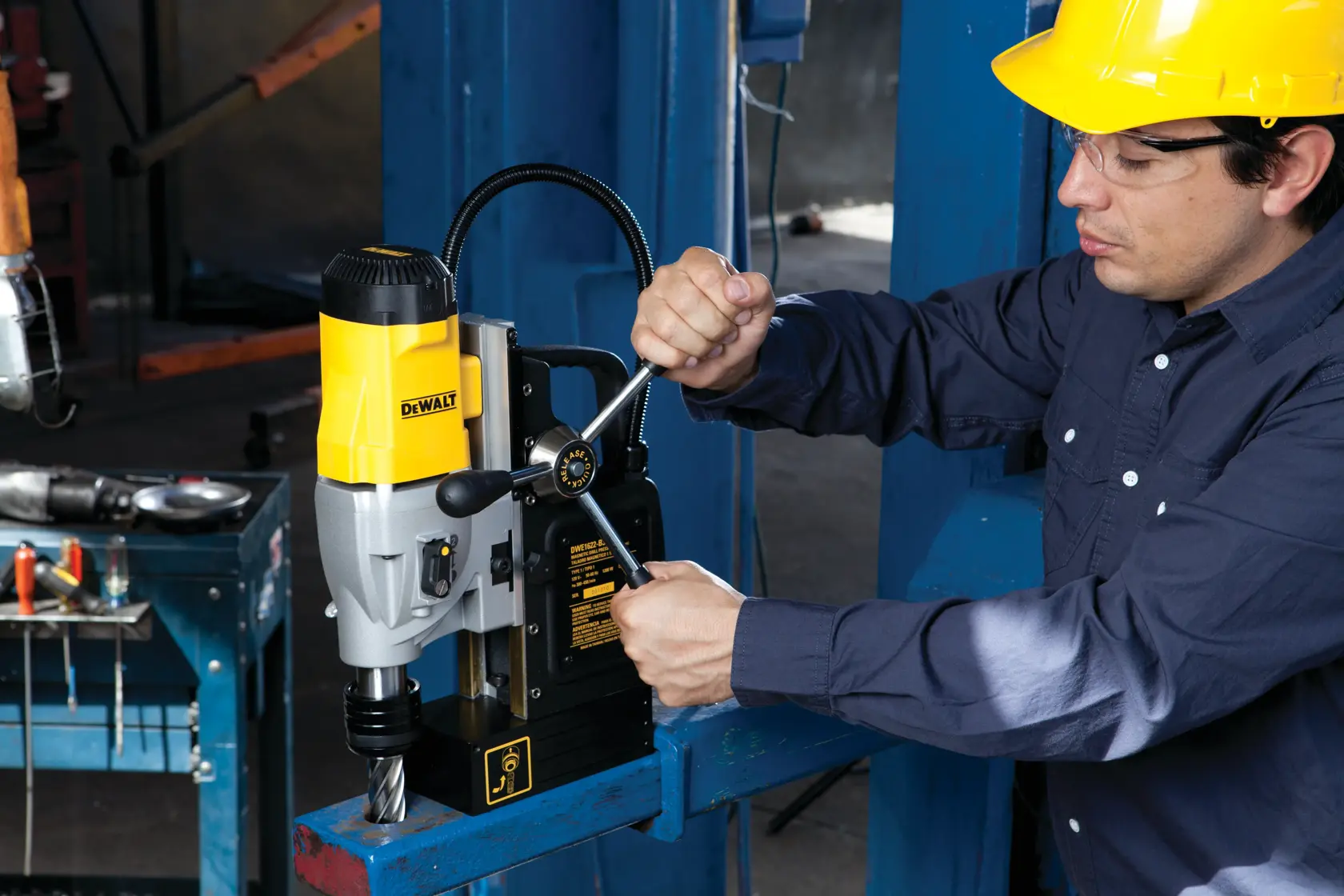 A person wearing a yellow hard hat operates a DEWALT magnetic drill press, model DWE1622K, mounted on a blue metal beam in an industrial workshop setting. The person's face is blurred for privacy.
