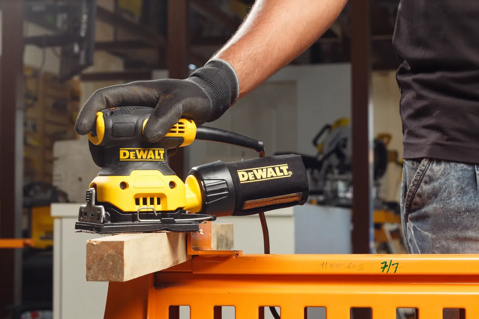 A gloved hand uses a DEWALT DWE6411 sander on a piece of wood in a workshop.