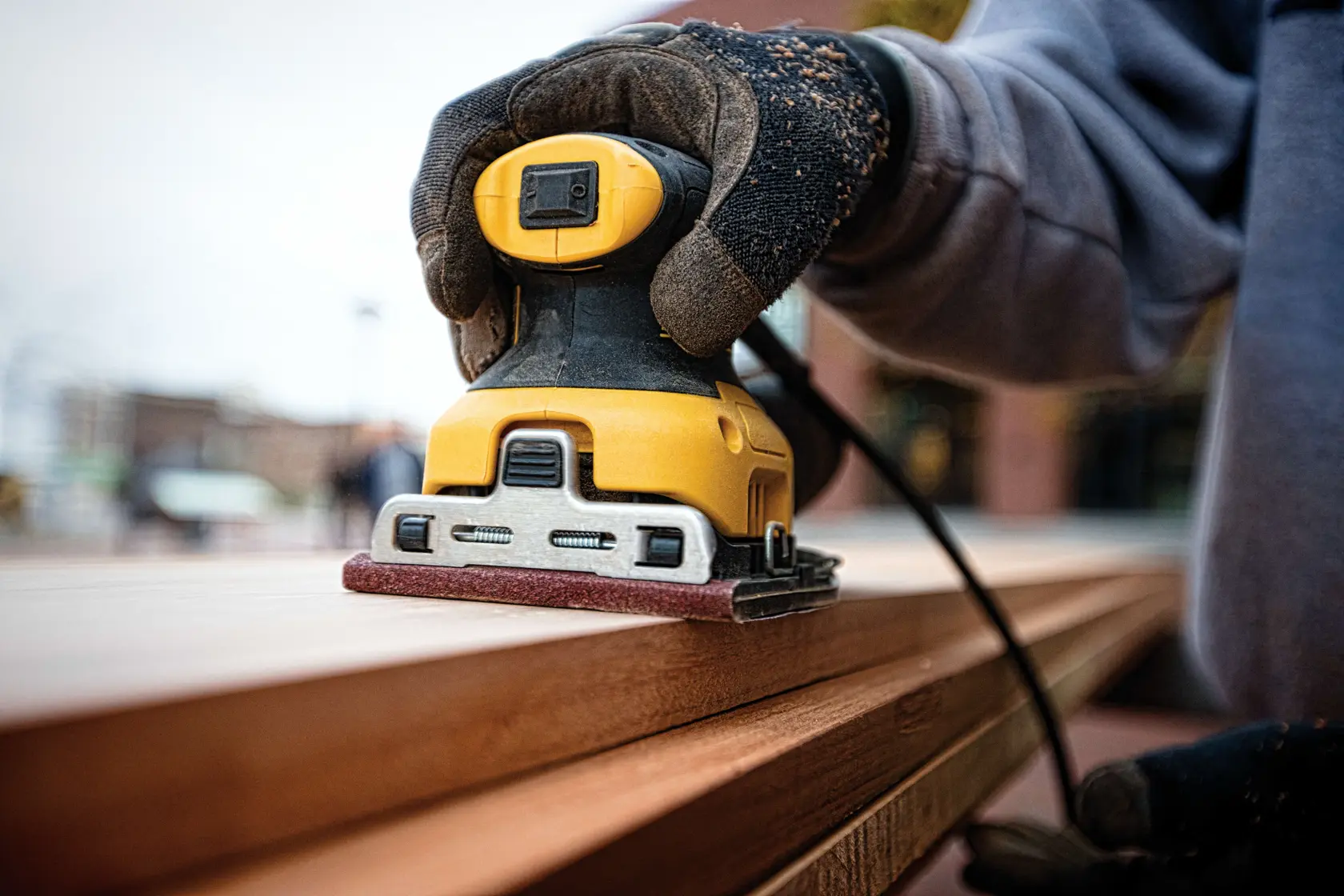 A close-up of a person wearing gloves using a yellow DEWALT electric sander to smooth the surface of a wooden plank.