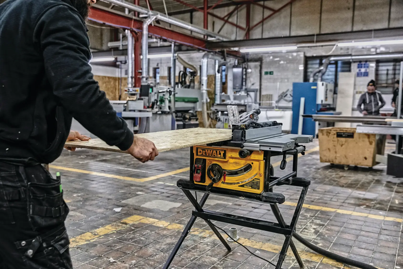 A person is using a yellow DEWALT table saw (model DWE7492SET) mounted on a black stand to cut a wooden board inside a workshop with industrial machines in the background.