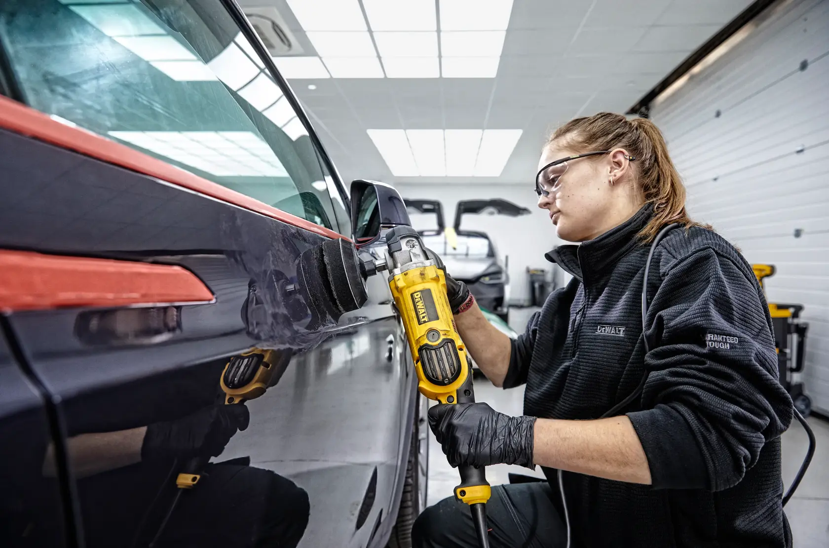 Person polishing a car door using a yellow DEWALT DWP849X tool in a workshop.
